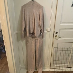 Beige stickad hoodie och byxor från Lindex - Mysig beige stickad hoodie och matchande byxor från Lindex. Perfekt för en avslappnad dag hemma eller ute. Hoodien har långa ärmar och ribbade muddar, medan byxorna har en bekväm resårmidja med snörning.