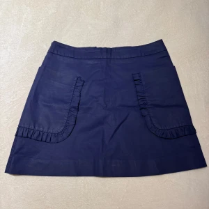 Mörkblå kjol från Mango - Snygg mörkblå kjol från Mango med volangdetaljer på fickorna. Kjolen har en dragkedja baktill och är perfekt för en stilren look. Tillverkad i ett bekvämt material som passar bra till olika tillfällen.