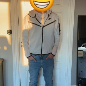 Nike Zip Vit - Snygg vit jacka från Nike med dragkedja och huva. Jackan har en stilren design med svarta detaljer och en ficka på ärmen. Perfekt för en sportig look.