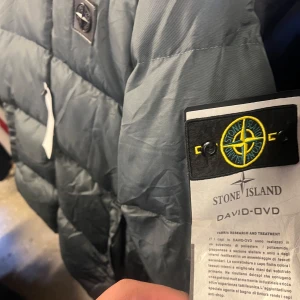 Grå dunjacka från Stone Island - Säljer en stilren grå dunjacka från Stone Island med dragkedja och ikonisk logga på ärmen. Jackan har en quiltad design och är perfekt för kyligare väder.