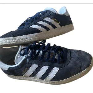 Adidas Gazelle i mörk blå och vit  - Snygga Adidas Gazelle sneakers i mörk blå mocka med vita detaljer och snörning. De har den klassiska tre-randiga designen och en vit sula. 