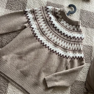 Beige stickad tröja från lager 157 - Mysig beige stickad tröja från 157 med ett vackert mönster i brunt och vitt runt halsen. Tröjan har långa ärmar och ribbstickade muddar vid ärmslut och nederkant. Perfekt för kyliga dagar! Storlek S