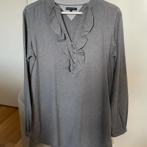 Ny Tommy Hilfiger blus  - Snygg och elegant Tommy Hilfiger blus i gråmelerad nyans.  Blusen har en v-ringning med en fin volangdetalj framtill och knappar. Långärmad med en avslappnad passform. Perfekt för både vardag och festligare tillfällen. •Helt ny, aldrig använd, bara tvättad •Nypris runt 1000 kr •Kommer från rökfritt och djurfritt hem •Storlek 6, passar till S
