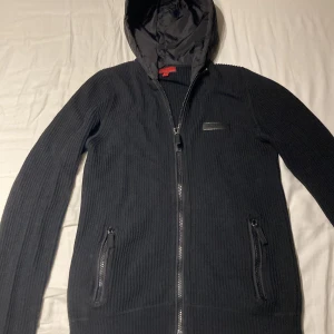 Svart stickad jacka från Burberry - Snygg svart stickad jacka från Burberry med dragkedja och huva. Jackan har en diskret logotyp på bröstet och praktiska fickor med dragkedja. Perfekt för kyliga dagar.