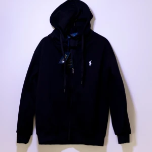 Svart hoodie från Ralph Lauren - Helt ny svart tröja från Ralph Lauren med dragkedja och klassisk logga på bröstet. Perfekt för en stilren och avslappnad look. Den har långa ärmar och justerbar huva med snören.