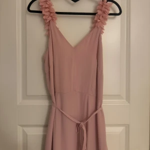 Ljusrosa långklänning med volangdetaljer - The Wardrobe - Elegant ljusrosa långklänning med volangdetaljer och knytband i midjan. Klänningen har breda axelband dekorerade med blommor och en v-ringad halslinje. Perfekt för sommarens festligheter. Perfekt för bal, bröllop eller midsommar. Skärpet är avtagbart och klänningen figursydd. 