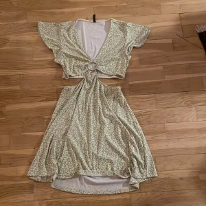 Blommig klänning från Divided - Säljer en charmig blommig klänning från Divided med en söt knutdetalj framtill. Klänningen har korta ärmar och en luftig passform, perfekt för varma dagar. Den har en ljus bas med ett diskret blommönster i grönt. Priset kan diskuteras.🫶