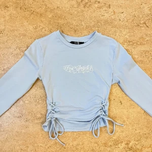 Ljusblå croppad topp med snörning - Snygg ljusblå croppad topp från Bershka med texten 'Los Angeles'. Toppen har långa ärmar och dekorativ snörning på sidorna som ger en unik look. Helt oanvänd (utanpris lapp), strl XS