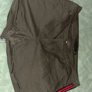 Olivgröna shorts från Amisu - Snygga olivgröna shorts från Amisu med röda och svarta ränder längs sidorna. De har en knapp och dragkedja framtill samt praktiska bakfickor. Perfekta för en avslappnad stil.