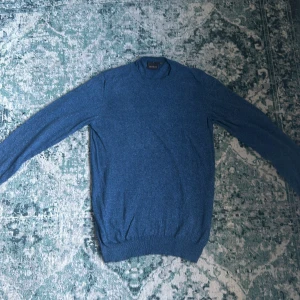 Blå tröja med unikt ullsymning från Oscar Jacobsson.  - Säljer en stilren blå tröja från Oscar Jacobson, tillverkad i Italien. Tröjan har en klassisk design med långa ärmar och ribbade muddar vid ärmslut och nederkant. Väldigt bra skick. Också mycket eftertraktad. Priset är inte hugget i sten.