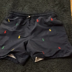 Svarta shorts från Ralph Lauren - Snygga svarta shorts från Ralph Lauren med broderade färgglada logotyper. Perfekta för en avslappnad stil. De har en bekväm passform med snörning i midjan.