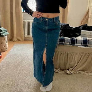 Blå jeanskjol från Bik Bok - Snygg, lång jeanskjol med en djup slits där fram i en blå/mörkblå färg och nyskick. Bra material och knappast använda. Kjolen har en högre midja men går att justera beroende på hur man tycker om att ha den. 