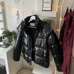 Svart pufferjacka från Calvin Klein - Snygg svart pufferjacka från Calvin Klein med glansig finish och huva. Jackan har en dragkedja framtill och är perfekt för kyligare dagar. Den har en klassisk pufferdesign som ger både stil och värme.