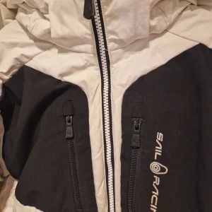 Vit och svart dunjacka från Sail Racing - Snygg och varm dunjacka från Sail Racing i vitt och svart. Jackan har en praktisk dragkedja framtill och två dragkedjefickor på bröstet. Perfekt för kyliga dagar med sin vadderade design och huva.