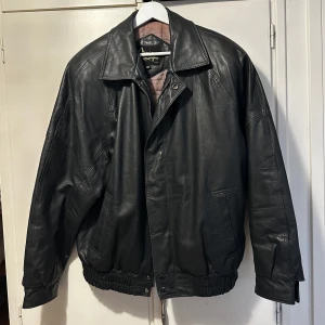 Svart skinnjacka från Manio Delfino - Säljer en klassisk svart läderjacka från manio Delfino. Jackan har en stilren design med dragkedja och knappar framtill. Perfekt för en cool och tidlös look.