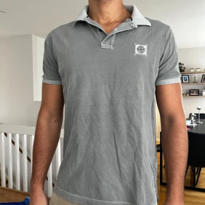 Grå pikétröja från Stone Island - grå pikétröja från Stone Island med korta ärmar och klassisk krage. Tröjan har en broderad logotyp på bröstet och knappar vid halsen. Perfekt för en avslappnad men snygg look. Storlek på tröja är XL men killen på bilden bär M i allt och är 180cm tror den har krympt 