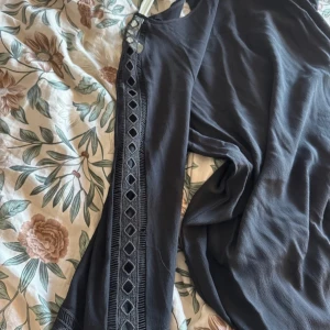 Svart klänning från KappAhl - Elegant svart klänning från KappAhl med dekorativa spetsdetaljer längs ärmarna. Klänningen har en klassisk design med trekvartsärmar och en rund halsringning. Perfekt för en stilren look.