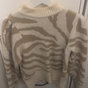 Beige och vit stickad tröja från Gina Tricot - Säljer en snygg stickad tröja från Gina Tricot i beige och vitt zebramönster. Tröjan har en hög ribbad krage och långa ärmar med puffiga axlar. Perfekt för att hålla sig varm och stilren under kyliga dagar.