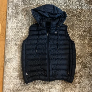 Blå dunväst från Campione del Garda - Snygg svart dunväst från Campione del Garda med dragkedja och huva. Perfekt för kyligare dagar och enkel att matcha med olika outfits. Nypris 1900kr och mitt pris är 499kr. Hör av dig vid några funderingar! 