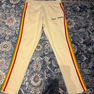 Beige byxor från Palm Angels med regnbågsränder - Snygga beige byxor från Palm Angels med färgglada ränder i rött, gult, grönt och blått längs sidorna. Byxorna har en elastisk midja och sidofickor. Perfekta för en avslappnad stil med en touch av färg.