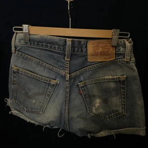 Blå jeansshorts från Levi Strauss & Co - Snygga blå lågmidjade jeansshorts från Levi's med slitna detaljer och råa kanter. Perfekta för en avslappnad stil. Klassisk femficksdesign och knappgylf. W 25 L 32
