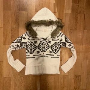 Vit stickad hoodie med mönster och fuskpäls - Mysig vit stickad hoodie med svart mönster och fuskpäls runt huvan. Perfekt för kyliga dagar med sin varma och bekväma design. Den har långa ärmar och en ribbad kant vid ärmslut och nederkant.