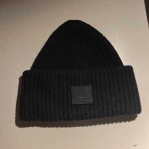 Svart ribbad mössa från Acne Studios - Snygg svart ribbad mössa från Acne Studios med en diskret logotyp framtill. Perfekt för att hålla värmen under kyliga dagar. Stilren design som passar till det mesta.
