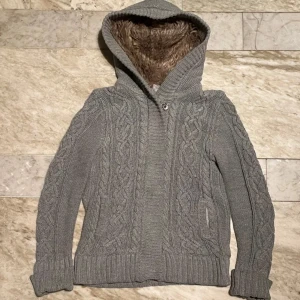 Grå stickad cardigan - Mysig grå stickad cardigan med kabelstickat mönster och mjukt foder i luvan. Perfekt för kyliga dagar med sin varma och bekväma design. Långa ärmar och ribbade muddar ger en skön passform.