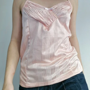 Rosa linne från ICHI - Säljer ett sött rosa linne från ICHI med tunna axelband och en liten rosett framtill. Linnet har ett elegant veckat mönster och är tillverkat i ett glansigt material som ger en lyxig känsla.