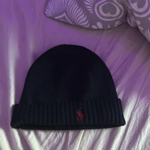 Svart mössa från Polo Ralph Lauren - Snygg svart mössa från Polo Ralph Lauren med ribbad kant och röd broderad logga framtill. Perfekt för att hålla värmen under kyliga dagar.