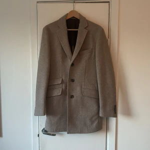 Beige rock från Brecos - Elegant beige rock från Brecos i en blandning av ull, polyamid och kashmir. Rocken har en klassisk design med tre knappar framtill och fickor med lock. Insidan är fodrad med ett stilrent rutigt mönster. Nypris 4500, mitt pris 2500. Använd en gång och är fodrad och varm.