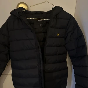 Svart dunjacka från Lyle & Scott - Säljer en svart dunjacka från Lyle & Scott med huva och dragkedja. Jackan har en klassisk design med det ikoniska logotypmärket på bröstet. Perfekt för kyliga dagar.