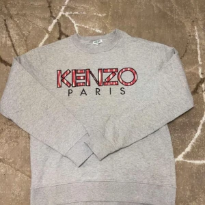 Grå sweatshirt från Kenzo - Säljer en stilren grå sweatshirt från Kenzo med det ikoniska logotyptrycket i rött och svart på bröstet. Tröjan har långa ärmar och en klassisk rund halsringning. Perfekt för en avslappnad look.