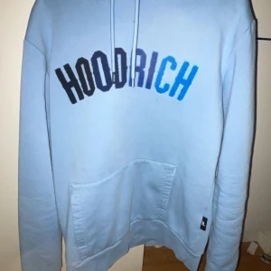 Ljusblå hoodie från Hoodrich - Säljer en snygg ljusblå hoodie från Hoodrich med text i svart och blått på bröstet. Den har en klassisk känguruficka och justerbar huva. Perfekt för en avslappnad stil.