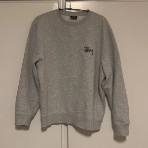 Grå sweatshirt från Stussy - Säljer en grå sweatshirt från Stussy med deras ikoniska logga tryckt både fram och bak. Tröjan har långa ärmar och en klassisk rund halsringning. Perfekt för en avslappnad stil.