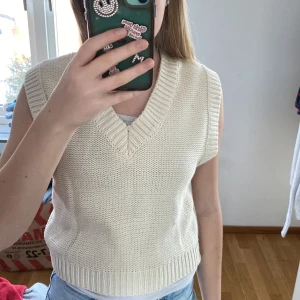 Beige stickad väst från Lindex - Säljer en stilren beige stickad väst från Lindex. Västen har en v-ringad design och ribbade kanter som ger en klassisk look. Perfekt för lager-på-lager-stil.