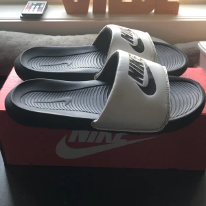 Svarta och vita Nike sandaler - Snygga och bekväma Nike sandaler i svart och vitt. Perfekta för sommardagar med en stilren design och Nike-logga på ovansidan. Sandalerna har en ergonomisk fotbädd för extra komfort. Köpt för 400 kr