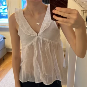 Vit ärmlös blus med volangdetaljer - Säljer en söt vit blus med volangdetaljer runt halsen. Blusen är ärmlös och har en knytning i ryggen. Helt ny, aldrig använd💕💕Storlek 170