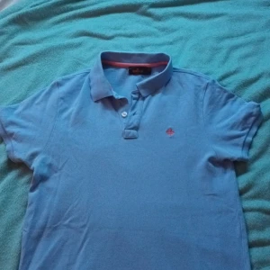 Blå pikétröja från Morris - Säljer en snygg blå pikétröja från Morris med korta ärmar och en liten broderad detalj på bröstet. Tröjan har en klassisk krage och knappar framtill. Perfekt för en avslappnad stil.