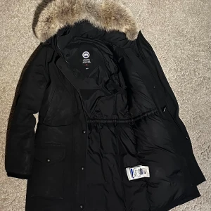 Svart parkajacka med pälsdetalj - Säljer en stilren svart parkajacka från Canada Goose med en lyxig pälsdetalj på huvan. Jackan har en lång design med dragkedja och knappar framtill, perfekt för kalla vinterdagar. Den är utrustad med flera praktiska fickor och har en varm och skyddande insida. Använd men i fint skick, finns ett litet 2mm reva under locket på ena fickan som lätt kan sys. Säljer den billigare pga det. 