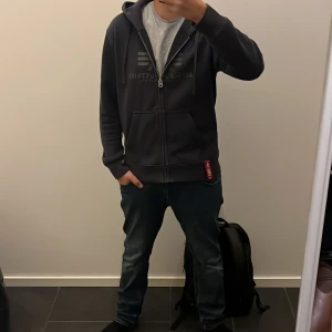 Svart hoodie från Alpha Industries - Säljer en svart hoodie från Alpha Industries med dragkedja och justerbar huva. Tröjan har en klassisk design med logotyp på bröstet och praktiska fickor framtill. Perfekt för en avslappnad stil. Storlek M men passar defenetivt S