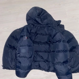 Svart pufferjacka från Tommy Hilfiger - Snygg mörkblå pufferjacka från Tommy Hilfiger med huva och broderad logga. Jackan har en quiltad design och är perfekt för kyligare väder. Den har långa ärmar och en klassisk passform.
