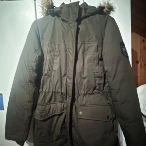 Olivgrön parkajacka med fuskpäls - Säljer en olivgrön parkajacka med huva och fuskpälsdetaljer. Jackan har långa ärmar och flera praktiska fickor framtill. Perfekt för kyliga dagar. Stor o rymlig i storleken. Använd ett fåtal gånger.