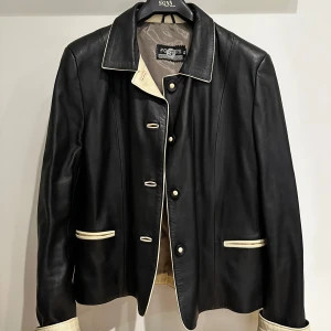 Svart skinnjacka Fosters Trading Company  - Snygg svart skinnjacka från Fosters Trading Company med kontrasterande vita detaljer. Jackan har knappar framtill och långa ärmar. Perfekt för en stilren look. Mjukt skinn, skick 9/10 ! Storlek 48, men skulle säga att den passar en S och vara lite oversized 🖤
