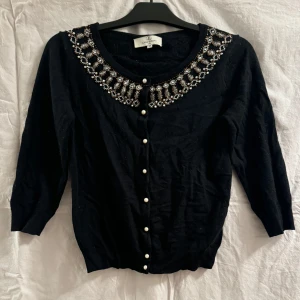 Svart kofta med pärldetaljer från Flash Woman - Elegant svart kofta från Flash Woman med vackra pärldetaljer runt halsringningen. Koftan har trekvartsärmar och knäpps med pärlknappar framtill. Perfekt för att ge en stilfull touch till din outfit.