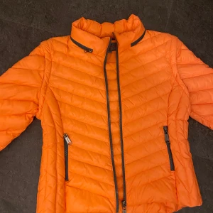 Orange pufferjacka - Snygg orange pufferjacka med dragkedja och hög krage. Jackan har långa ärmar och praktiska fickor med dragkedjor. Kan ta bort ärmar så det blir en väst samt huva. Vårjacka storlek 44 men passar mig som har S i vanliga fall.
