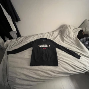 Marin blå tröja från Morris - Säljer en marin blå tröja från Morris med text och en brittisk flagga på framsidan. Tröjan har långa ärmar och en klassisk rund halsringning. Perfekt för en stilren och avslappnad look.