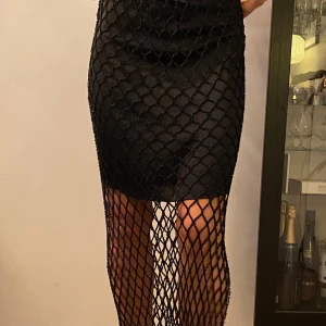 Svart glittrig kjol med nät och pärlor  - Snygg svart kjol med ett glittrigt nätmönster som ger en unik look. Kjolen är tight och har en elegant längd som passar perfekt för speciella tillfällen. Detaljerna i nätet ger en extra touch av glamour.