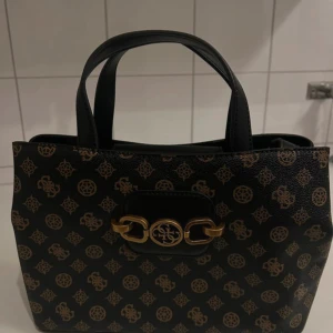 Svart handväska med mönster - Snygg svart handväska med ett elegant mönster i brunt. Väskan har dubbla handtag och en praktisk dragkedja på baksidan. Insidan är rymlig med flera fack och stängs med en knapp. Perfekt för att hålla ordning på dina saker. Aldrig används 