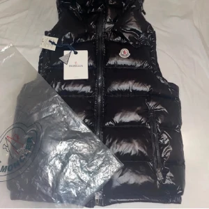 Svart dunväst från Moncler - Snygg svart dunväst från Moncler med glansig yta och dragkedja framtill. Västen har Monclers logotyp på bröstet och en unik etikett med serietidningsmotiv på insidan. Perfekt för kyligare dagar. Använd att fåtal gånger men inte varje dag, den har inga skador och är i 10/10 skick!